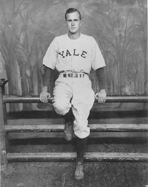 George Bush standing in front of a "Yale Fence" in his baseball uniform
