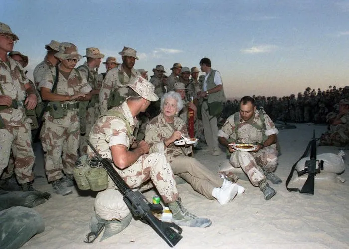 First Lady Barbara Bush sits in the sand eating Thanksgiving Dinner with Marines of the First Marine Division in Saudi Arabia