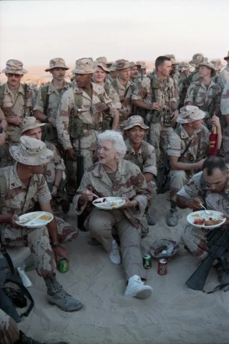 First Lady Barbara Bush sits in the sand eating Thanksgiving Dinner with Marines of the First Marine Division in Saudi Arabia